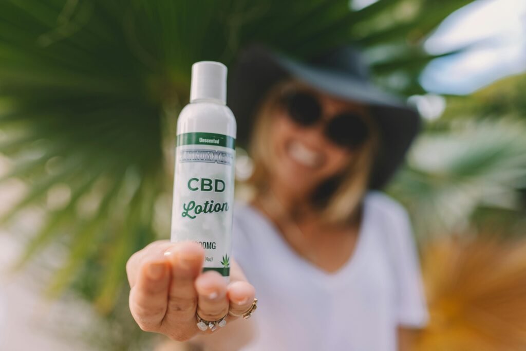 A woman in a hat smiling and holding a CBD lotion bottle with a blurred palm background.